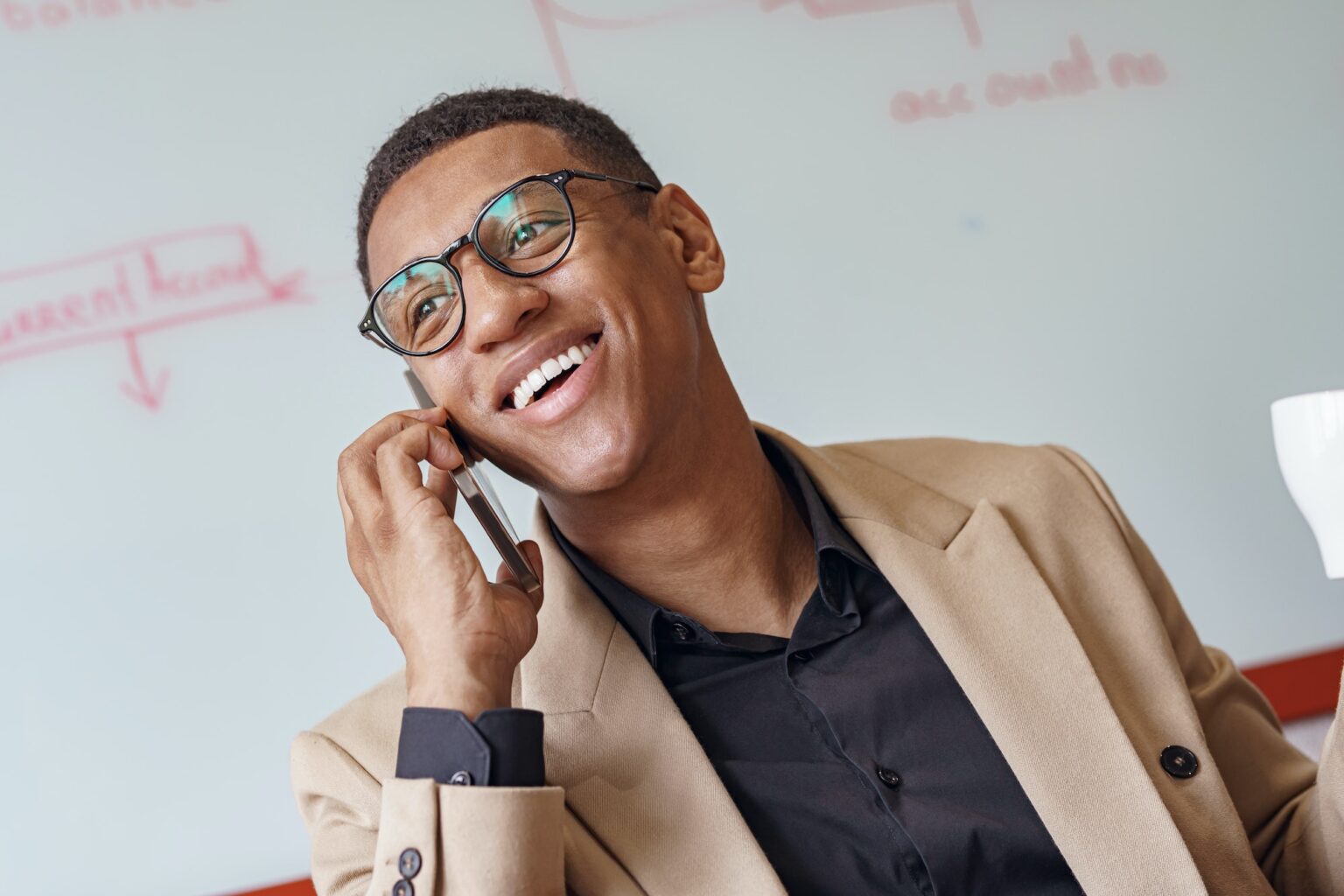 Smiling african businessman in glasses talking phone sitting in cozy coworking and looking away